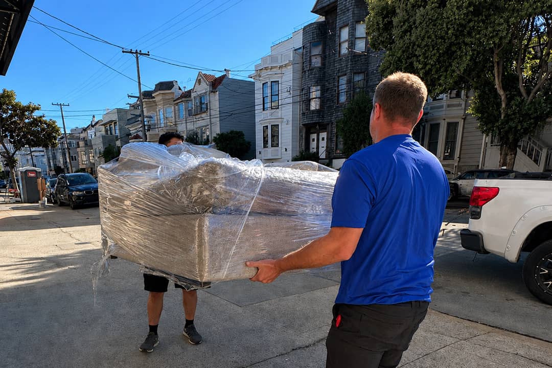 Professional movers carrying a plastic-wrapped couch across a street.